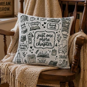 May include: A white square throw pillow featuring black line art illustrations of books, a reading chair, a candle, and socks. The central text reads, "just one more chapter," surrounded by phrases like "book lover," "currently reading," and "book club."