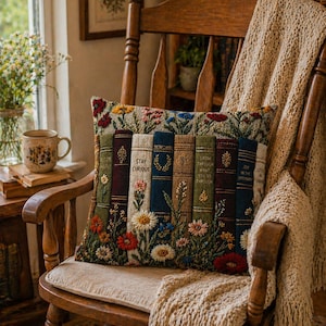 May include: A decorative pillow with a floral and book design. The pillow features embroidered images of books with floral accents and the text "Stay Curious" and "Grow Through What You Go Through". The pillow is on a wooden rocking chair.