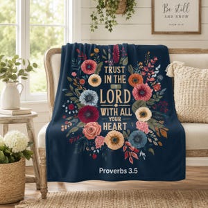 May include: A navy blue throw blanket featuring a floral wreath design surrounding the text "TRUST IN THE LORD WITH ALL YOUR HEART" in gold lettering, with the citation "Proverbs 3.5" printed in white at the bottom of the fabric.