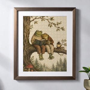 May include: A framed jigsaw puzzle featuring an illustration of two green frogs sitting on a tree branch. One frog wears a beige jumper and reads a book, while the other wears a brown jacket and holds a mug next to a small glowing lantern.