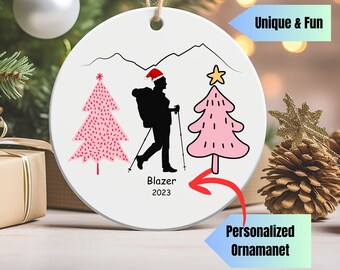 May include: White ceramic Christmas ornament with a silhouette of a hiker wearing a Santa hat, carrying a backpack and hiking poles, with two pink Christmas trees on either side. The ornament has the text "Blazer 2023" at the bottom.