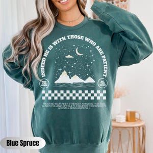 K&ouml;nnte beinhalten: Gr&uuml;ner Pullover mit einem wei&szlig;en Grafikdesign mit einer Sichelmond, Sternen, Bergen und dem Text "Indeed he is with those who are patient". Die Grafik enth&auml;lt auch den Text "See the good" und "Be the good" in kleinerer Schrift. Der Text "Trusting your inner strength, knowing that ease always follows difficulty, enduring the hardships and still being grateful" ist unter der Grafik gedruckt.
