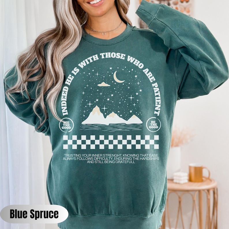 K&ouml;nnte beinhalten: Gr&uuml;ner Pullover mit einem wei&szlig;en Grafikdesign mit einer Sichelmond, Sternen, Bergen und dem Text "Indeed he is with those who are patient". Die Grafik enth&auml;lt auch den Text "See the good" und "Be the good" in kleinerer Schrift. Der Text "Trusting your inner strength, knowing that ease always follows difficulty, enduring the hardships and still being grateful" ist unter der Grafik gedruckt.