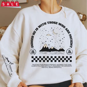 K&ouml;nnte beinhalten: Wei&szlig;er Pullover mit einem schwarzen Grafikdesign, das eine Sichelmond, Sterne, Berge und den Text "Indeed He is with those who are patient." zeigt. Die Grafik enth&auml;lt auch zwei kleine Quadrate mit dem Text "See the good" und "Be the good". Der Text unter der Grafik lautet "Trusting your inner strength, knowing that ease always follows difficulty, enduring the hardships and still being grateful."