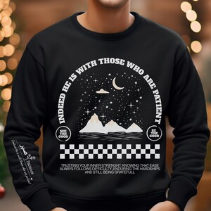 K&ouml;nnte beinhalten: Schwarzer Pullover mit einem wei&szlig;en Grafikdesign, das eine Sichelmond, Sterne, Berge und den Text "Indeed he is with those who are patient." zeigt. Die Grafik ist von einem karierten Muster umgeben. Der Text "See the good" und "Be the good" sind ebenfalls im Design enthalten. Der Text unter der Grafik lautet "Trusting your inner strength, knowing that ease always follows difficulty, enduring the hardships and still being grateful."