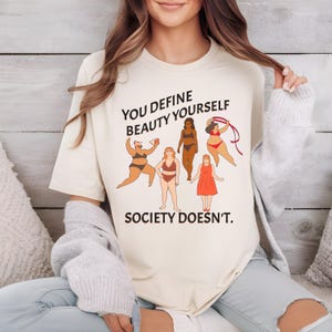 K&ouml;nnte beinhalten: Ein beiges T-Shirt mit dem Text "YOU DEFINE BEAUTY YOURSELF SOCIETY DOESN'T." und einer Illustration von f&uuml;nf Frauen mit unterschiedlichen K&ouml;rperformen.