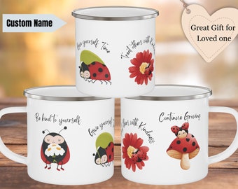 May include: White enamel mugs with silver rims, featuring ladybug and flower illustrations. Each mug has a different inspirational phrase, such as "Give yourself time" and "Be kind to yourself." The mugs are a great gift.