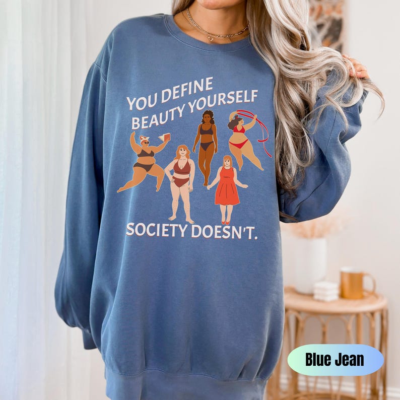 K&ouml;nnte beinhalten: Blauer Pullover mit einem Grafikdruck von f&uuml;nf Frauen mit unterschiedlichen K&ouml;rperformen und Hautt&ouml;nen. Der Text "You define beauty yourself. Society doesn't." ist auf dem Pullover aufgedruckt.