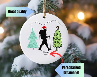 May include: White ceramic Christmas ornament with a hiker in a red Santa hat walking through a snowy mountain landscape with two green Christmas trees. The ornament is personalized with the name "James" and the year "2023".
