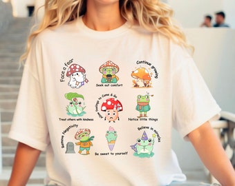 May include: White t-shirt with a colorful graphic design featuring frogs and mushrooms. The design includes phrases like "Face a fear", "Seek out comfort", "Continue growing", "Treat others with kindness", "Remove negativity", "Be sweet to yourself", "Allow feelings to come & go", "Notice little things", and "Believe in miracles".