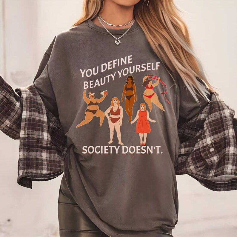 K&ouml;nnte beinhalten: Ein graues Langarmshirt mit dem Text "You define beauty yourself, society doesn't." und einer Illustration von f&uuml;nf Frauen mit unterschiedlichen K&ouml;rperformen und Hautt&ouml;nen.