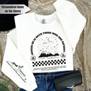 K&ouml;nnte beinhalten: Wei&szlig;er Pullover mit einem schwarzen Grafikdesign mit einer Sichelmond, Sternen, Bergen und dem Text "Indeed He is with those who are patient". Die Grafik ist von einem schwarzen Schachbrettmuster umgeben. Der &Auml;rmel tr&auml;gt den Text "Patience" und ein kleines Herz.