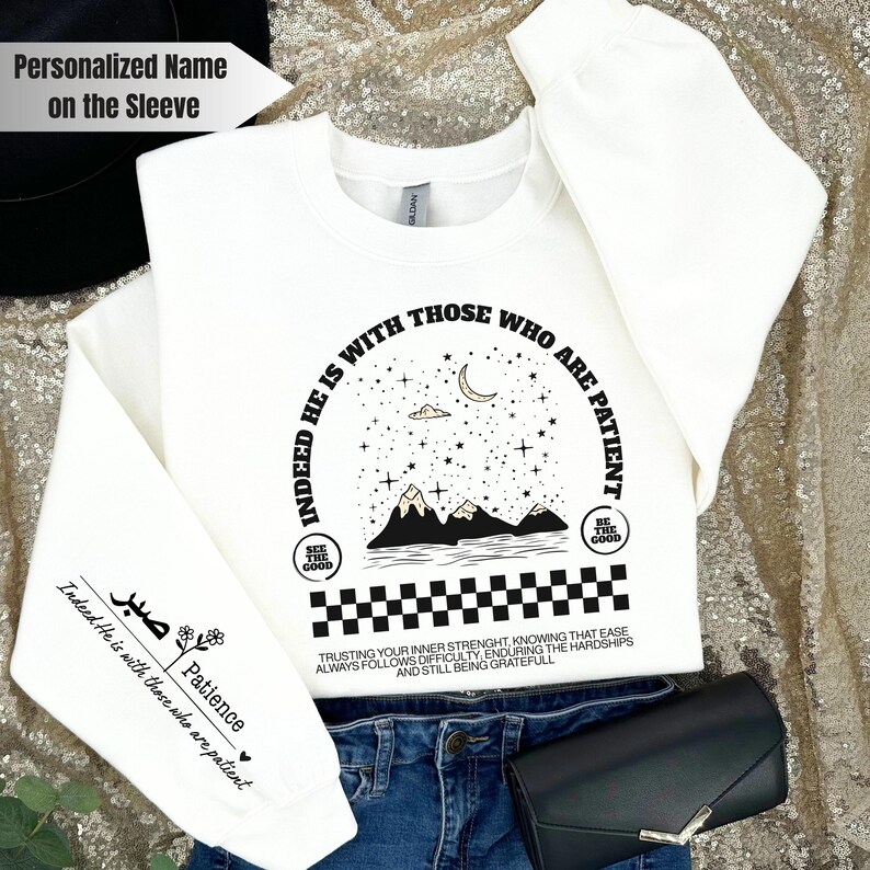 K&ouml;nnte beinhalten: Wei&szlig;er Pullover mit einem schwarzen Grafikdesign mit einer Sichelmond, Sternen, Bergen und dem Text "Indeed He is with those who are patient". Die Grafik ist von einem schwarzen Schachbrettmuster umgeben. Der &Auml;rmel tr&auml;gt den Text "Patience" und ein kleines Herz.