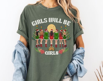 May include: Moss green t-shirt with the text "GIRLS WILL BE GIRLS" above a graphic of six girls in Christmas outfits. The graphic includes Christmas trees, a full moon, and poinsettias. The shirt has rolled-up sleeves.