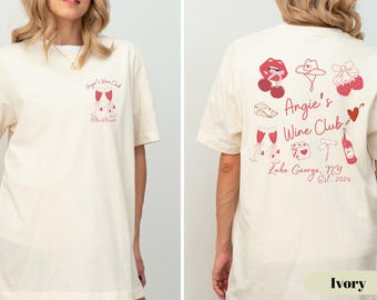 以下が含まれることがあります： アイボリー色のTシャツ。前面には「Angie's Wine Club」という赤いグラフィックが、背面には「Angie's Wine Club, Lake George, NY Est. 2024」という赤いグラフィックがプリントされています。