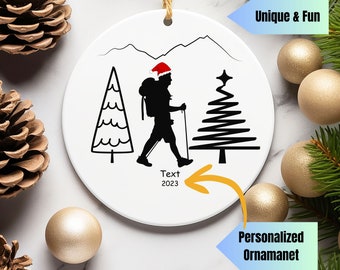 May include: White ceramic Christmas ornament with a black silhouette of a hiker wearing a Santa hat, walking between two stylized Christmas trees. The ornament has the text "Text 2023" on it.