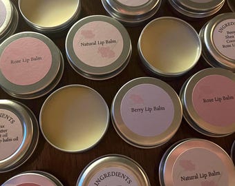 May include: A collection of round metal tins containing lip balm. Some tins are closed with labels reading Berry Lip Balm, Rose Lip Balm, Natural Lip Balm, or Ingredients including beeswax, shea butter, and coconut oil. Others are open.