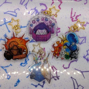Può includere: Una collezione di portachiavi in acrilico con chiusure dorate a forma di stella. Ogni portachiavi presenta un design diverso, tra cui dadi, fiamme e testo come "Dungeon Master", "Artificer" e "Sorcerer". I portachiavi sono su uno sfondo con un motivo a costellazioni.