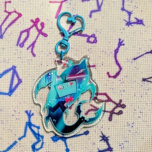 Op de afbeelding: Een heldere acryl bedel met een cartoonfiguur in teal en paars. De bedel heeft een teal hartvormige sluiting. Het karakter heeft een tv-vormig hoofd, een rode vlinderdas en een zwarte staart. De achtergrond is een lichte stof met paarse en blauwe borduursels.