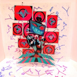 Może przedstawiać: Kolorowy stojak akrylowy przedstawiający postać z kreskówki z głową w postaci ekranu telewizora i szerokim, uśmiechniętym uśmiechem. Postać jest osadzona na tle czerwonych i czarnych kwadratów z turkusowymi akcentami. Podstawa ma turkusowe i różowe paski.