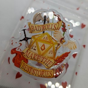Könnte beinhalten: Ein klarer Resin-Abzeichen mit Fantasy-Thema. Das Abzeichen zeigt einen gelb-goldenen D20-Würfel, ein Banner mit der Aufschrift "BAD ROLLS MAKE THE BEST STORIES" und Illustrationen eines Schwertes, einer Axt und einer geflügelten Kreatur. Das Abzeichen ist in einer transparenten, herzförmigen Tasche eingeschlossen.