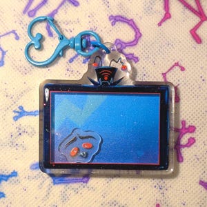Könnte beinhalten: Ein blauer und schwarzer Acryl-Schlüsselanhänger mit einem Cartoon-Fernsehdesign mit blauem Bildschirm und einem kleinen, durchsichtigen Cartoon-Charakter. Der Schlüsselanhänger hat einen blauen Verschluss.