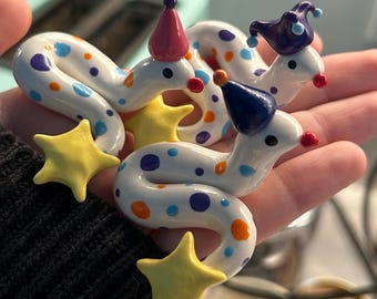 May include: Three white ceramic snake figurines with colorful polka dots, each wearing a small party hat in red, dark blue, or purple. Each snake features a bright yellow star attached to its body and a small red nose on its face.