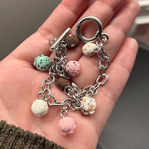May include: A silver-toned metal chain bracelet featuring multiple miniature charms shaped like scoops of ice cream in various colors including mint green, chocolate brown, pink, white, and speckled varieties, secured with a toggle clasp closure.