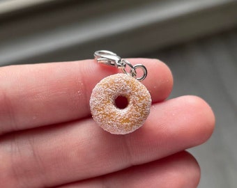 May include: A miniature donut charm with a silver clasp. The donut is light brown with a white, sugary coating. The charm is designed to be attached to a bracelet or necklace.