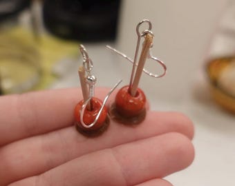 May include: Handmade earrings with red beads and wooden accents. The earrings have silver-coloured ear wires and sit on a small, brown base. The design resembles miniature toffee apples. The earrings are a unique accessory.