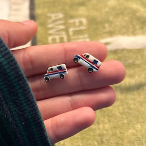 Könnte beinhalten: Miniatur-Ohrringe in Form eines weißen Posttransporters mit roten und blauen Streifen. Die Ohrringe haben schwarze Räder und sind vor einem verschwommenen Hintergrund platziert. Die Ohrringe sind klein und detailliert.