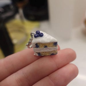 Könnte beinhalten: Ein weißer Kuchen-Anhänger mit blauem Zuckerguss und Blaubeeren darauf. Der Kuchen ist ein Stück Kuchen und ist ein Anhänger für eine Halskette oder ein Armband.