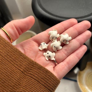 Op de afbeelding: Vijf kleine, witte, spookvormige beeldjes met gladde, glanzende oppervlakken en twee kleine zwarte stippen als ogen. Elk spookje heeft een afgerond hoofd en gedrapeerde, vloeiende randen, wat zorgt voor een uniform uiterlijk van de stukken.