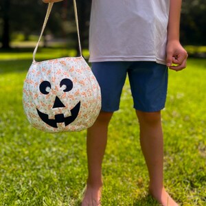 May include: A round fabric pumpkin bucket featuring a white background with a small orange floral pattern. The bucket has a black felt jack-o'-lantern face with two circular eyes and a wide, toothy grin, and includes a single fabric handle.