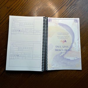May include: A book with a purple and blue watercolor design on the cover. The title of the book is "Once Upon a Broken Heart" by Stephanie Garber. The book cover also features a quote: "Heroes don't get happy endings, they give them to other people."