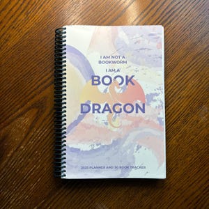 May include: A spiral bound planner and book tracker with a watercolor background. The cover features a dragon with the text "I am not a bookworm I am a book dragon" and "2025 Planner and 50 Book Tracker".