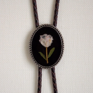 May include: A bolo tie featuring a dark brown braided leather cord and an oval-shaped pendant. The pendant has a silver-toned twisted metal border surrounding a black background with a single pressed white flower and green leaves in the center.
