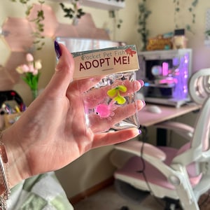 Puede incluir: Un paquete de plástico transparente que contiene dos pequeños peces de juguete de colores. El paquete tiene una cabecera de cartón con el texto "Pocket Pet Fish" y "ADOPT ME!". Un pez es rosa, el otro es amarillo y verde.