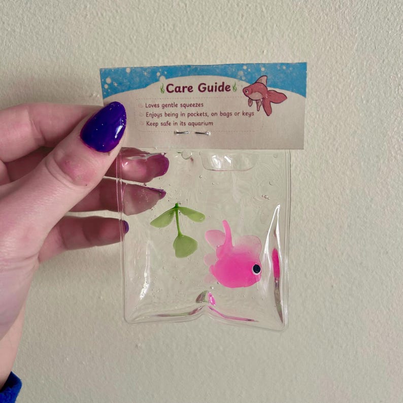 May include: A clear plastic bag containing a small pink fish figurine and a green plant sprig. The attached header card features a blue and white design with the text Care Guide, listing instructions: Loves gentle squeezes, Enjoys being in pockets, on bags or keys, Keep safe in its aquarium.