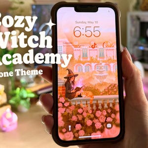 May include: A smartphone screen shows a "Cozy Witch Academy Phone Theme" with a witch reading a book. The screen displays a pink and orange illustration of a witch, cat, and roses. The phone is in a brown case.