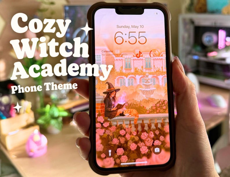 May include: A smartphone screen shows a "Cozy Witch Academy Phone Theme" with a witch reading a book. The screen displays a pink and orange illustration of a witch, cat, and roses. The phone is in a brown case.