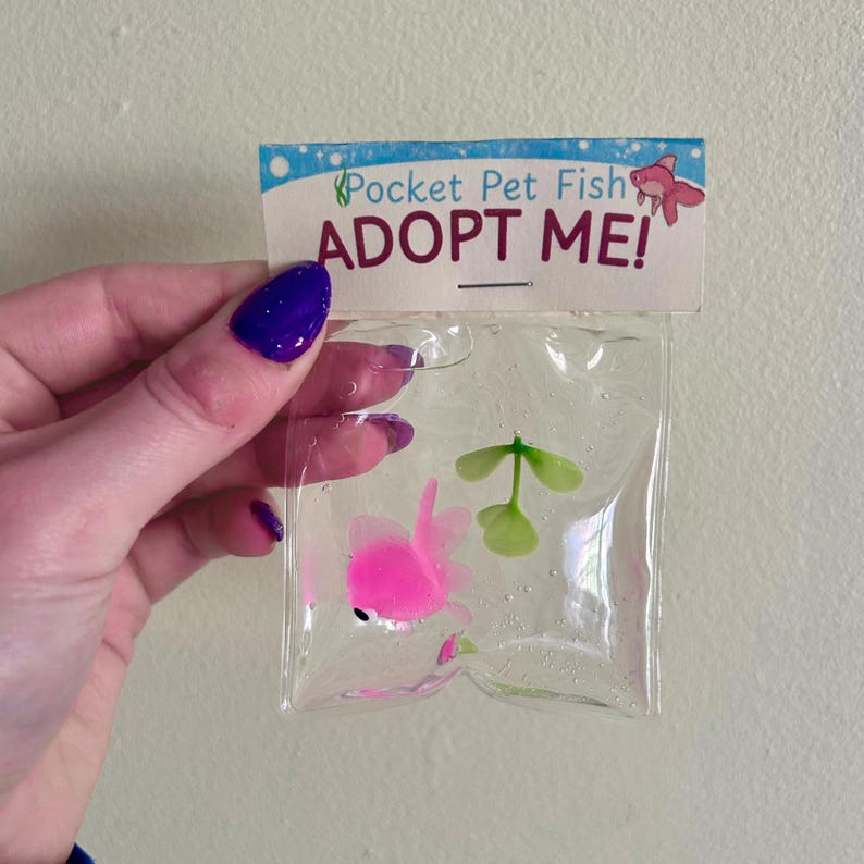 May include: A clear plastic bag containing a small pink fish figurine and a green plant piece. The top of the bag features a white cardboard header with blue bubble graphics, the text Pocket Pet Fish, and the bold red text ADOPT ME!.
