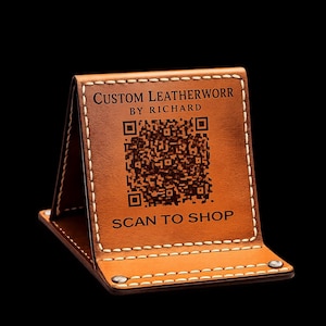May include: A tan leather stand with white stitching, featuring the text CUSTOM LEATHERWORR BY RICHARD, a central QR code, and the text SCAN TO SHOP. The stand is folded into a triangular shape and secured with silver-toned metal rivets at the base.