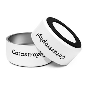May include: Two white pet bowls with stainless steel interiors. One bowl is upright, the other is tilted, revealing the interior. The word "Catastrophe!" is printed on the side of the bowls in black, playful font. The bowls have a black rubber ring.