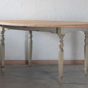 Op de afbeelding: Een witte ovale tafel met een houten blad en gedraaide poten. De tafel heeft een verweerde witte verfbehandeling en is perfect voor een boerderij of cottage stijl huis.