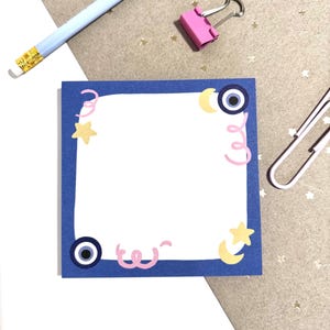 Puede incluir: Una nota adhesiva cuadrada azul con un interior blanco y un borde azul oscuro. Las esquinas están decoradas con espirales rosas, estrellas amarillas, lunas crecientes y símbolos azules de mal de ojo. También se ven un lápiz azul claro, una pinza rosa y un clip plateado.