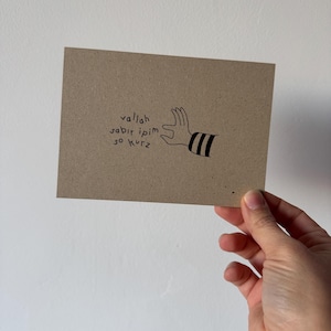 Könnte beinhalten: Eine braune Karte mit dem Text "vallah sabir ipim so kurz" und einer Handillustration mit schwarzen Streifen. Die Karte wird vor einem weißen Hintergrund gehalten.