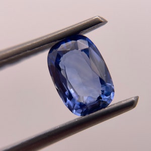 Puede incluir: Una piedra preciosa ovalada de color azul oscuro sostenida por unas pinzas. La piedra preciosa está facetada y tiene un alto pulido.