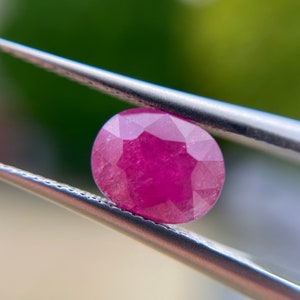 May include: A close-up of a round, faceted ruby gemstone held by tweezers. The ruby is a deep pink color and has a high level of brilliance.