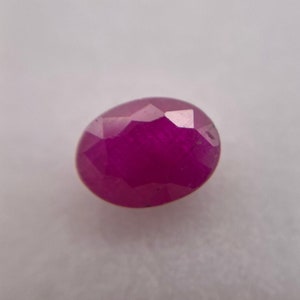 May include: A deep pink oval-shaped ruby gemstone with a faceted cut.